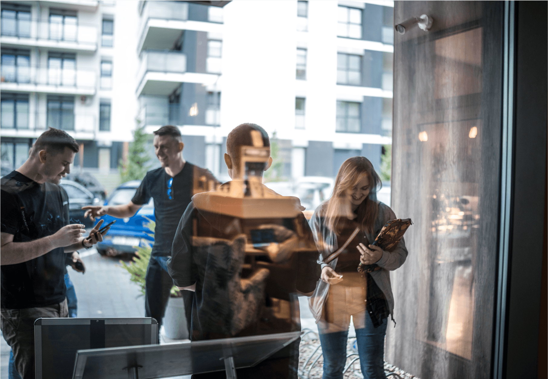 Group outside, seen through a window, smiling with phones and tablets, reflecting indoors.