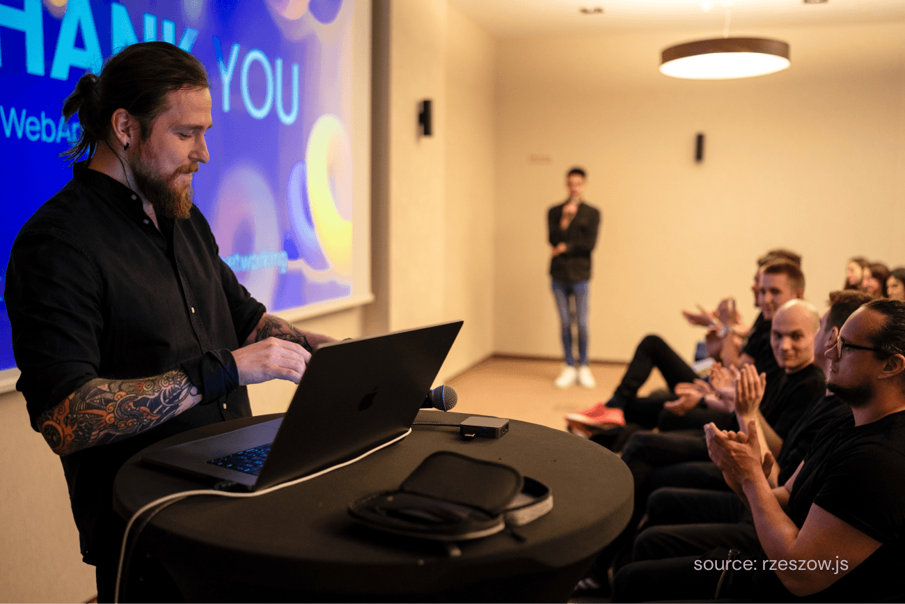 Man with a laptop giving a conference, people in the background clapping.