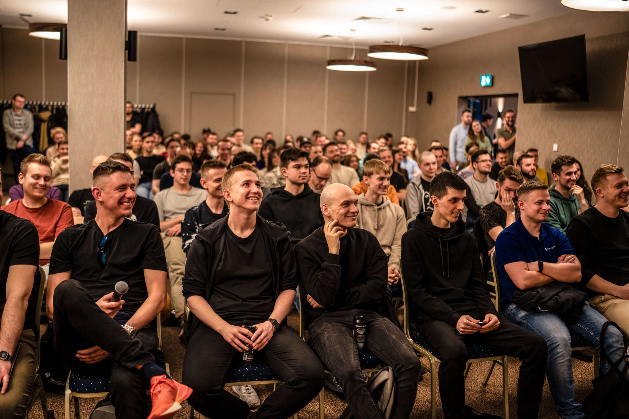 Large group in a conference room, smiling and engaging with each other, some sitting, some standing.