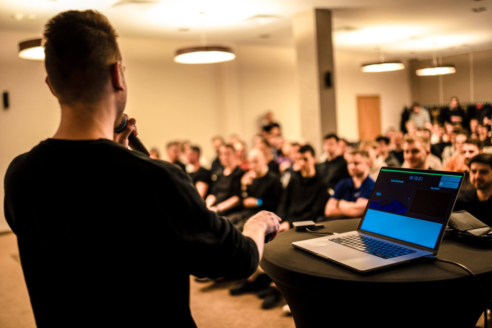 Presenter speaking into a microphone with slides on a laptop beside them, in front of an audience.