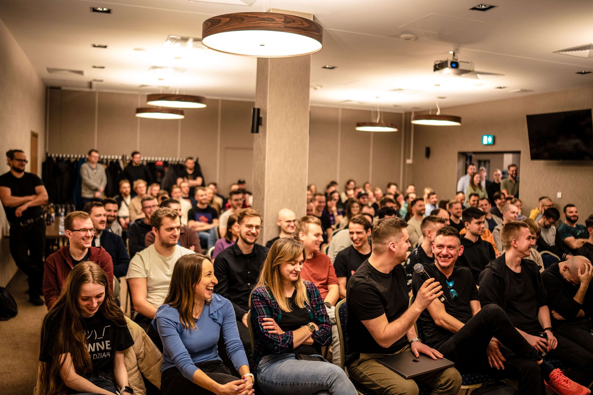 Large group in a conference room, many smiling, laughing, and holding microphones.