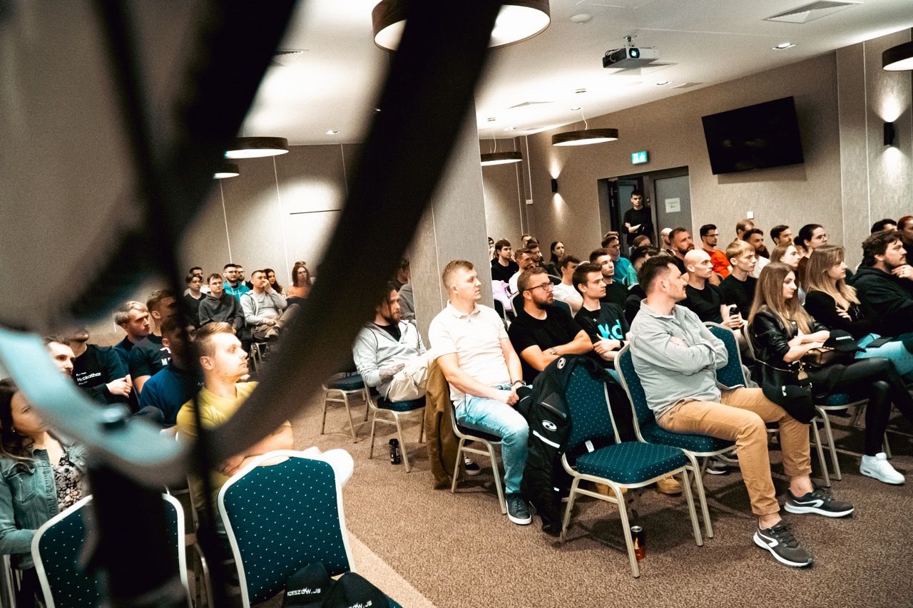 Audience in rows, attentively facing the front, with some objects partially obscuring the view.