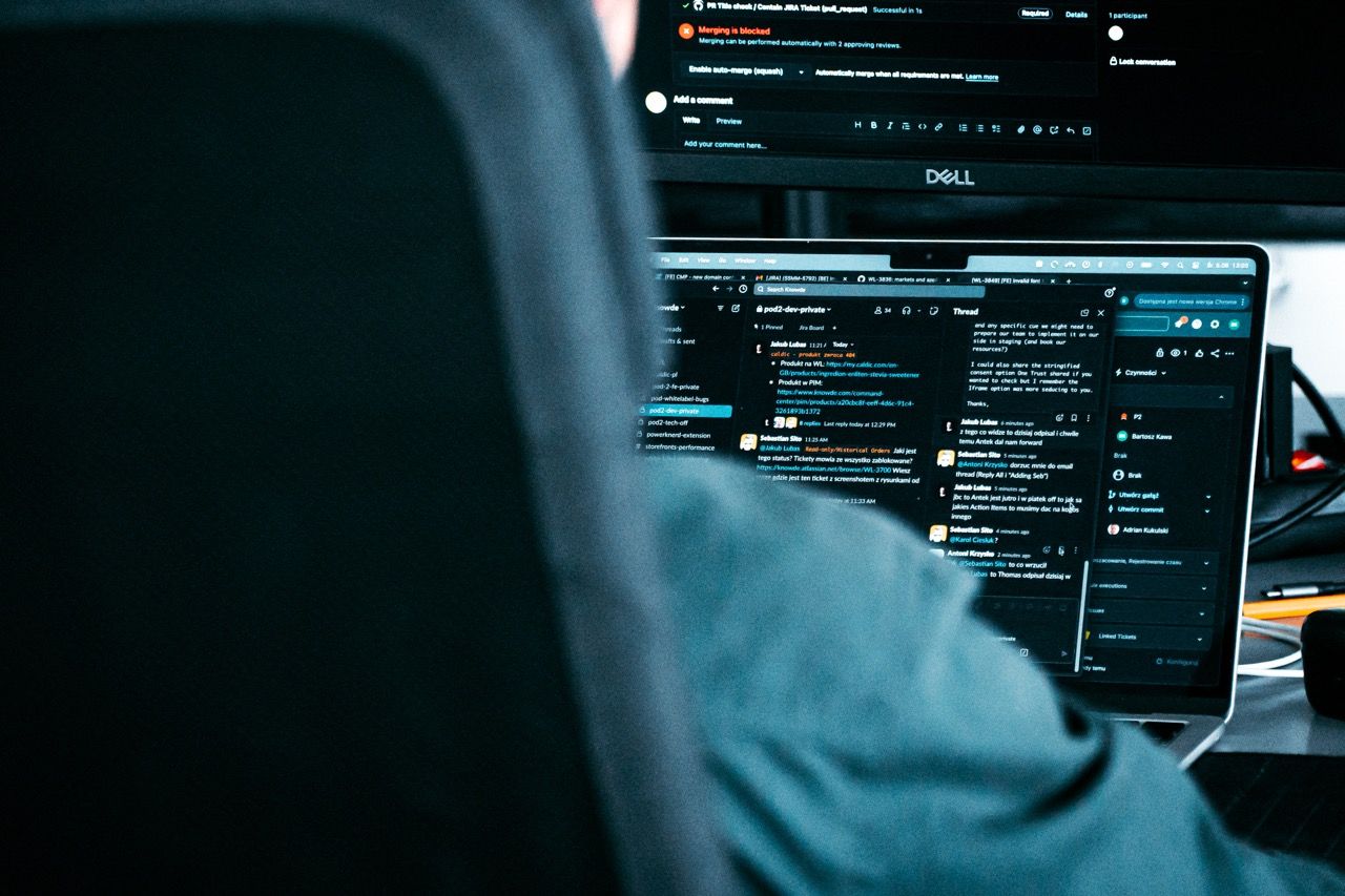 Close-up of a laptop screen with coding and communication apps, person working in background.