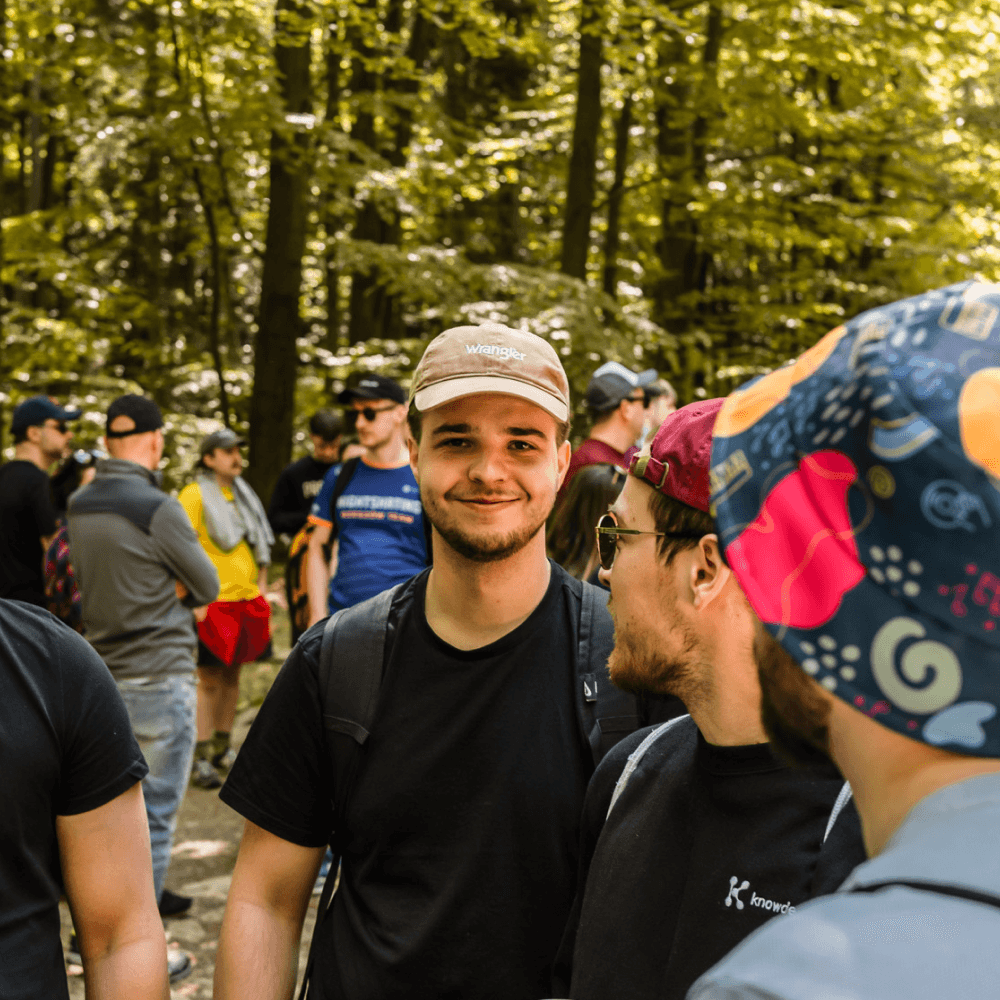 Man in a cap smiling, standing in a forest with a group of people in the background