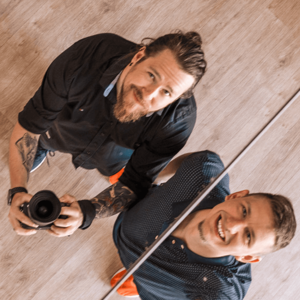 Two men looking up at the camera, one holding a camera, standing on a wooden floor