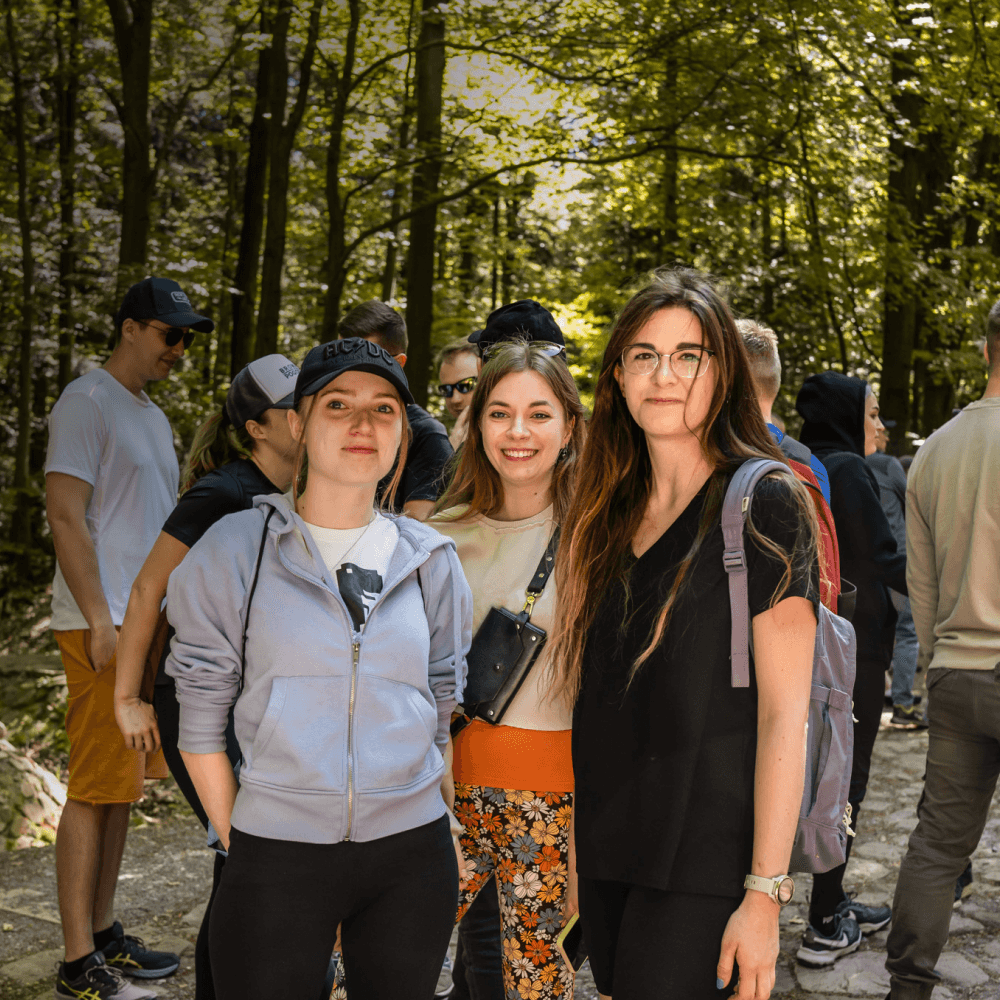 Women standing and smiling in the foreground of a wooded area, with other people in the background